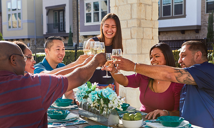 Six people sit at a table outdoors and hold up wine glasses