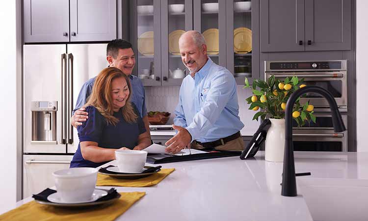 A David Weekley Homes Warranty Manager stands in a kitchen a man and a woman, all three people are looking at a notebook on the counter