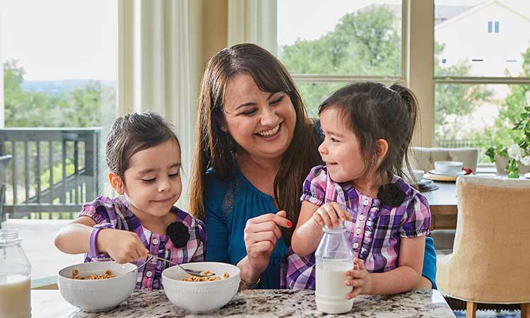 A woman sits at a kitchen counter with two children, the children are eating cereal with milk