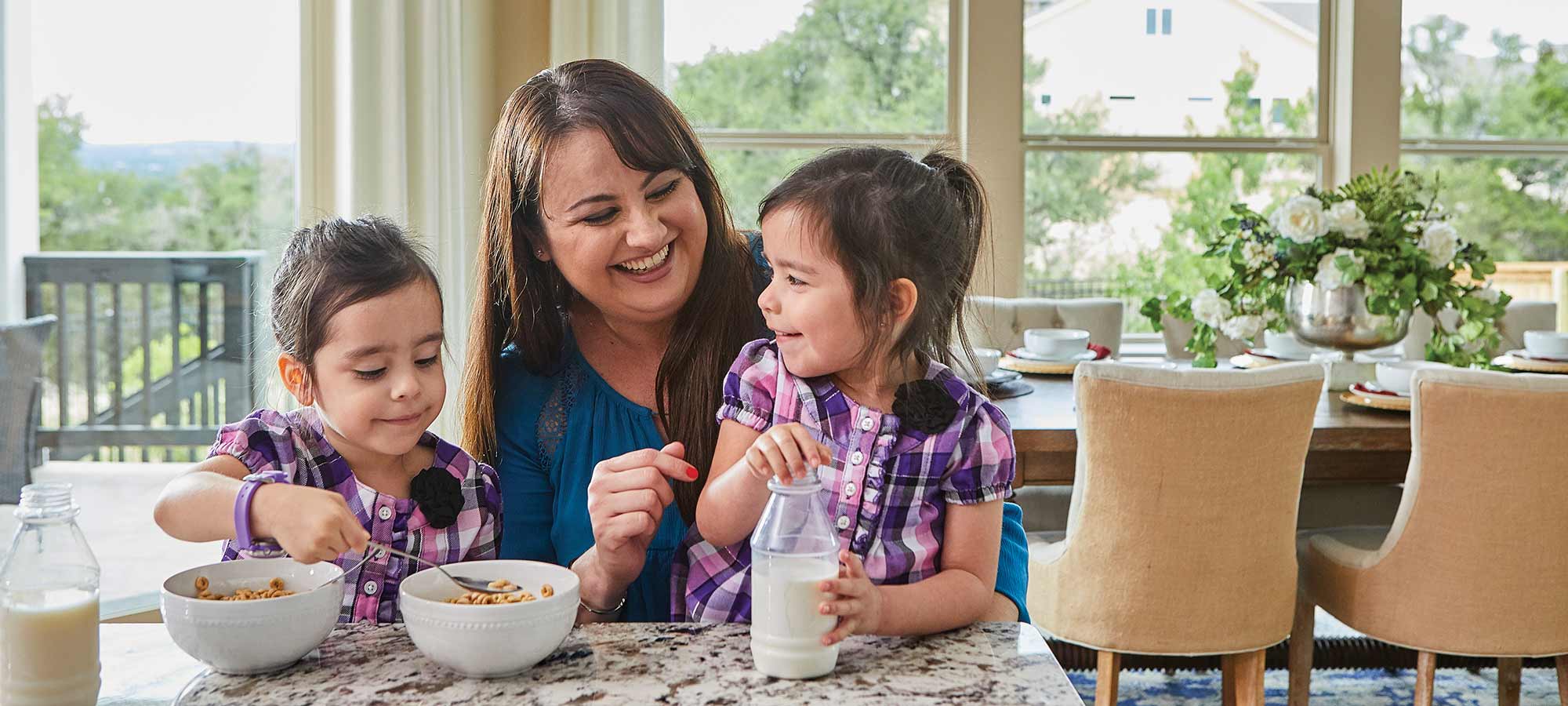 a woman sits at a kitchen counter with two young children, the children are eating cereal out of bowls