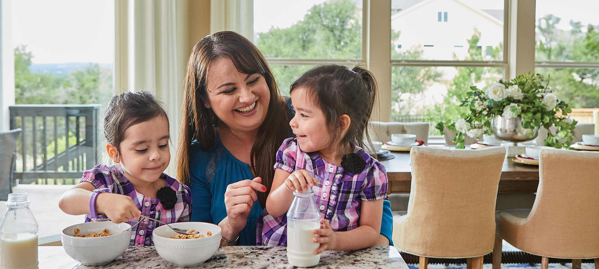 a woman sits at a kitchen counter with two young children, the children are eating cereal and milk