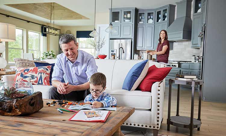 A child sits at a coffee table with coloring pages and crayons, a man sits on a couch smiling while a woman is standing in the kitchen, she is smiling as well