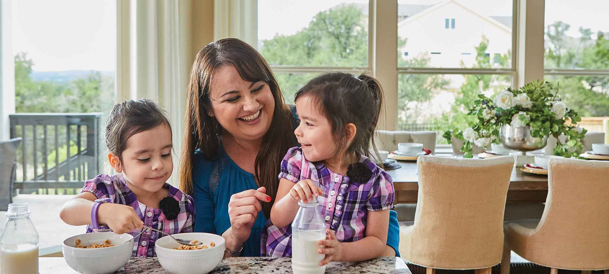 A woman sits at a kitchen counter with two young girls, they are two bowls of cereal on the counter and jars of milk