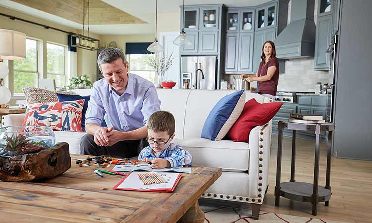 a child sits at a coffee table with crayolas and a coloring book, a man and woman stand nearby smiling