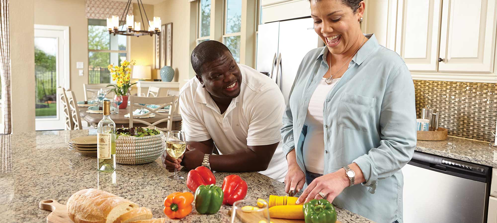 a man and a woman stand at a kitchen counter, the woman is cutting vegetables and the man is leaning on the counter and smiling