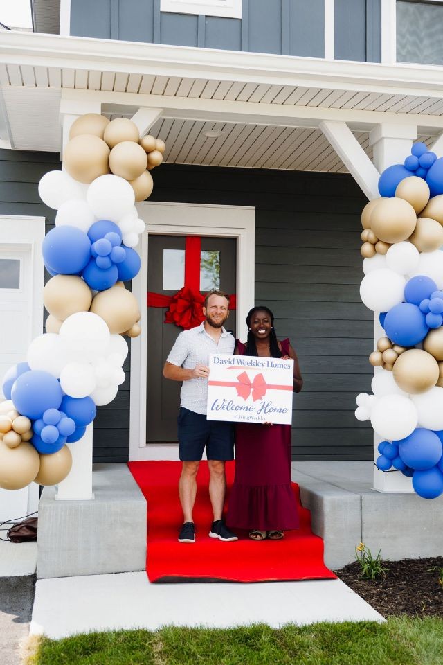 a man and a woman standing on a front porch holding up a Welcome Home sign