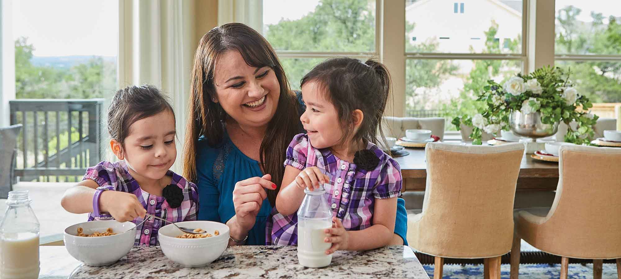 A woman sits at a kitchen counter with two, young children. The children are eating cereal.