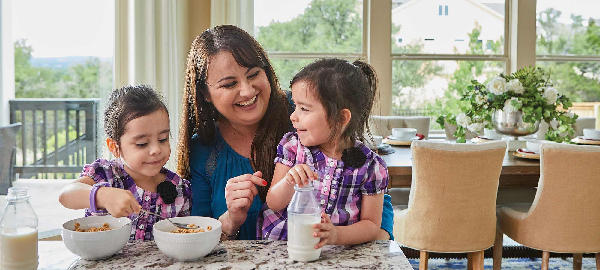 a woman sits at a kitchen counter with two young children, the children are eating cereal with milk