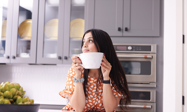 A woman leans over a kitchen counter while holding a cup