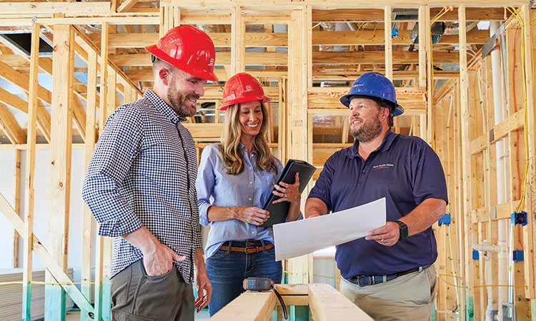 A David Weekley Homes Personal Builder stands in a home that is under construction with a man and a woman, all three people are wearing hard hats and looking at a large piece of paper