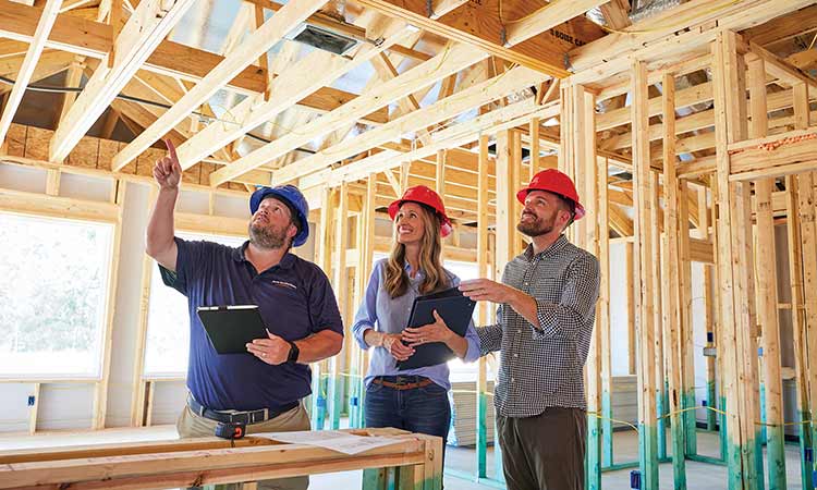 A David Weekley Homes Personal Builder stands in a home under construction with a man and a woman. All three people are wearing hardhats
