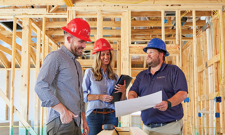 A David Weekley Homes Personal Builder stands in a home under construction with a man and a woman. All three people are wearing hard hats