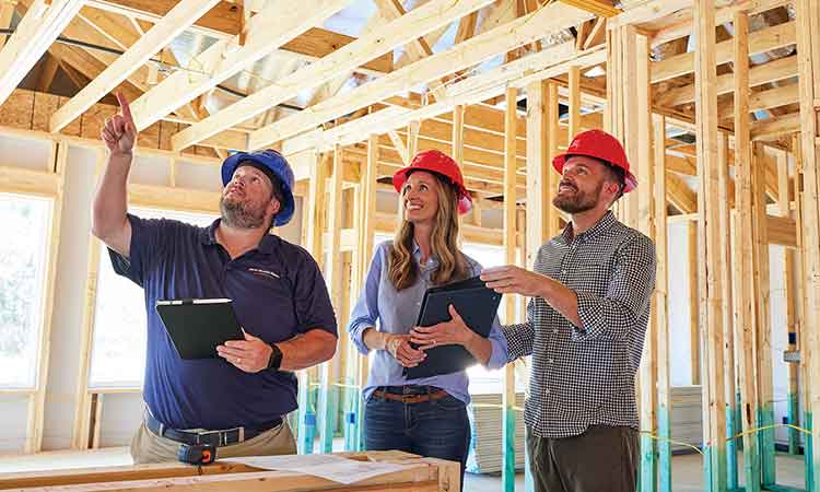 David Weekley Homes Personal Builder is standing in a home that is under construction with a man and a woman. The builder is pointing up towards the ceiling. All three people are wearing hardhats