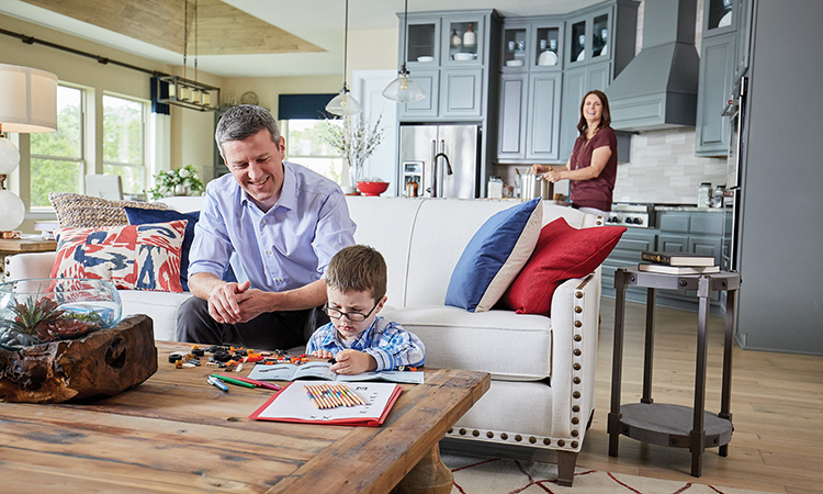 a child sits at a coffee table drawing on a piece of paper, a map sits nearby on a couch and woman is standing in a kitchen